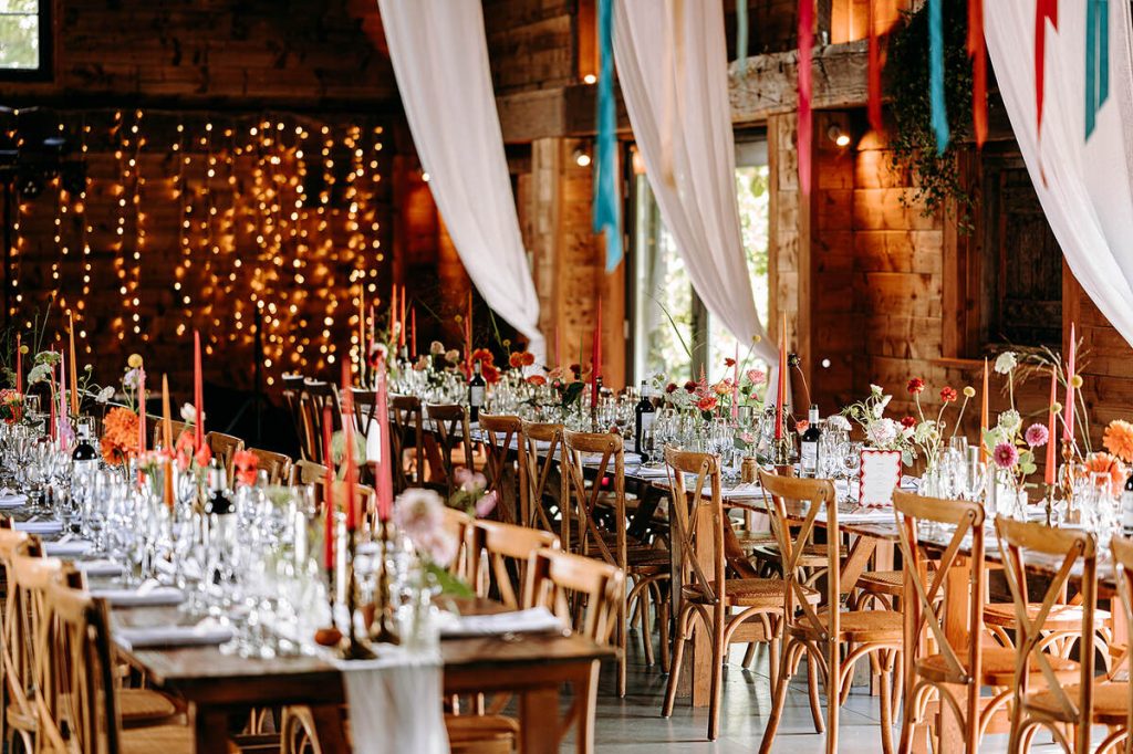 A wedding barn set up with décor and styling with a colourful, festival vibe. Long wooden tables are laden with flowers, crockery, cutlery and glassware with wooden chairs and colourful streamer banners hanging overhead.