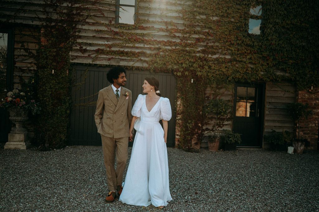 A bride and groom stroll arm in arm outside their barn wedding venue whilst gazing lovingly at one another. The groom wears a tan brown suit whilst his wife wears a plunging neck white linen wedding dress with puff sleeves.