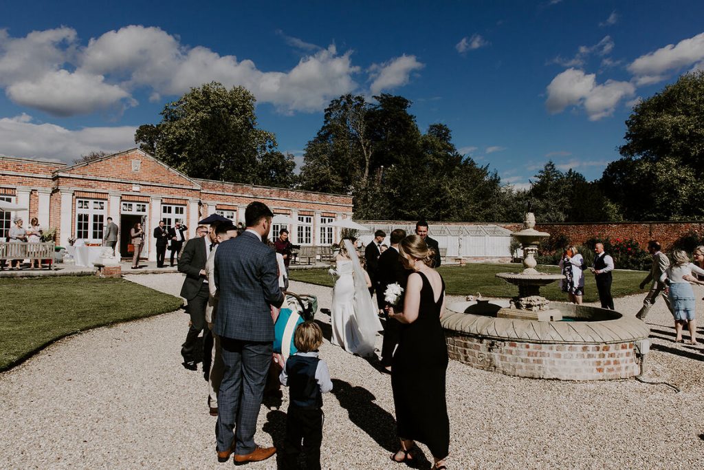 Guests mill around a walled garden with blue skies overhead during a wedding reception. A working fountain takes centre stage of the beautifully manicured gardens around which the guests chat and relax.