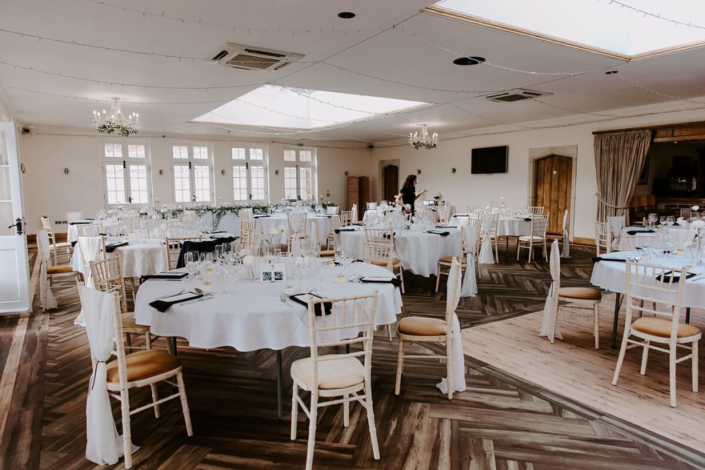 Minimalist wedding styling inside an orangery with round tables clothed in white and simple black napkins on each table. A long white clothed top table is lined with simple green foliage and candles.
