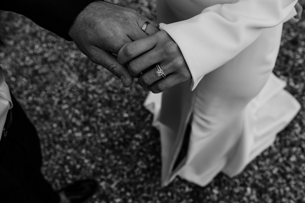 A bride and groom hold hands showing off their engagement rings and wedding bands featuring a lot of diamonds.