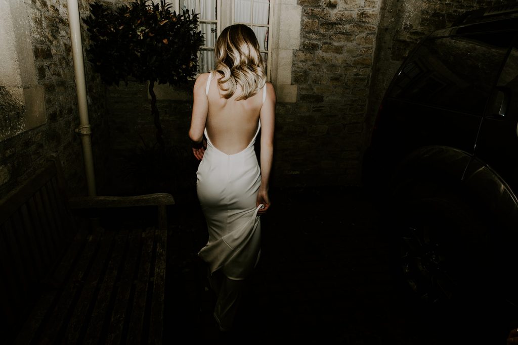 A bride walks away from the camera as the flash goes off, illuminating her in the evening darkness. Her mid length blonde hair is loosely curled and she wears a slinky, satin slip type dress with a sensuous plunging open back.