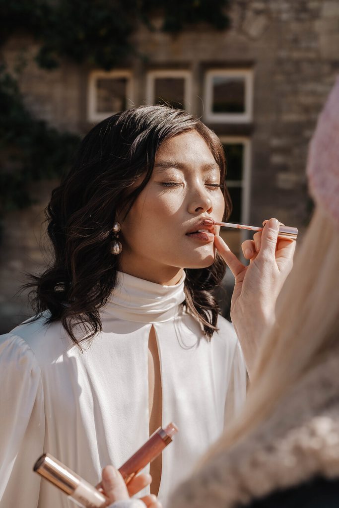 A bride wearing a high neck wedding dress closes her eyes as her bridal makeup artist applies more lipstick to her lips.