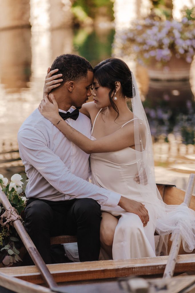 A newlywed couple sit inside a small rowing boat on a small boating lake wearing their wedding clothes. They caress one another in an intimate, tender moment. The bride wraps her hands and arms gently around her husband's face and neck as they gently rest their foreheads on one another's.