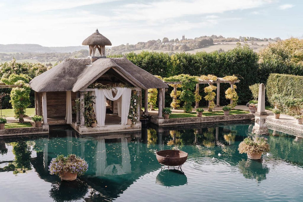 A wedding venue in the countryside with a draped cloth ceremony area looks out over a small boating lake. In the distance, rolling green countryside hills can be seen as a backdrop.