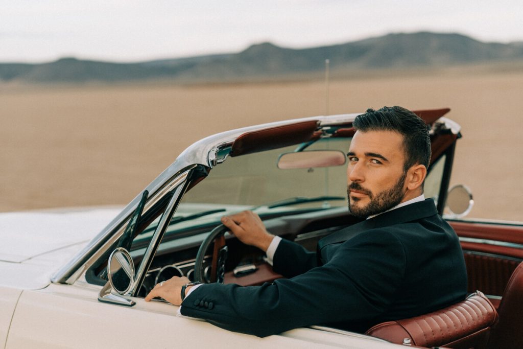 A groom sits in the driving seat of a classic convertible Mustang, holding the steering wheel with his right hand whilst he looks over his left shoulder with a smouldering look.