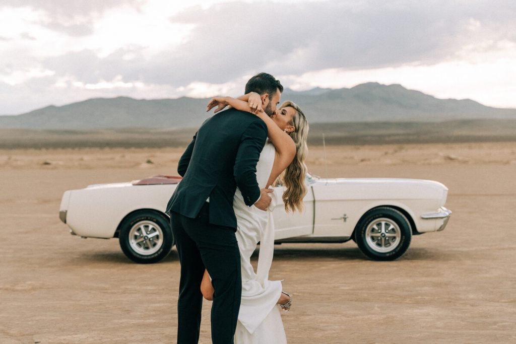 A bride and groom share a kiss in the desert in front of their parked Mustang classic convertible car. The bride throws her arm around her grooms neck and raises one leg back in the air.