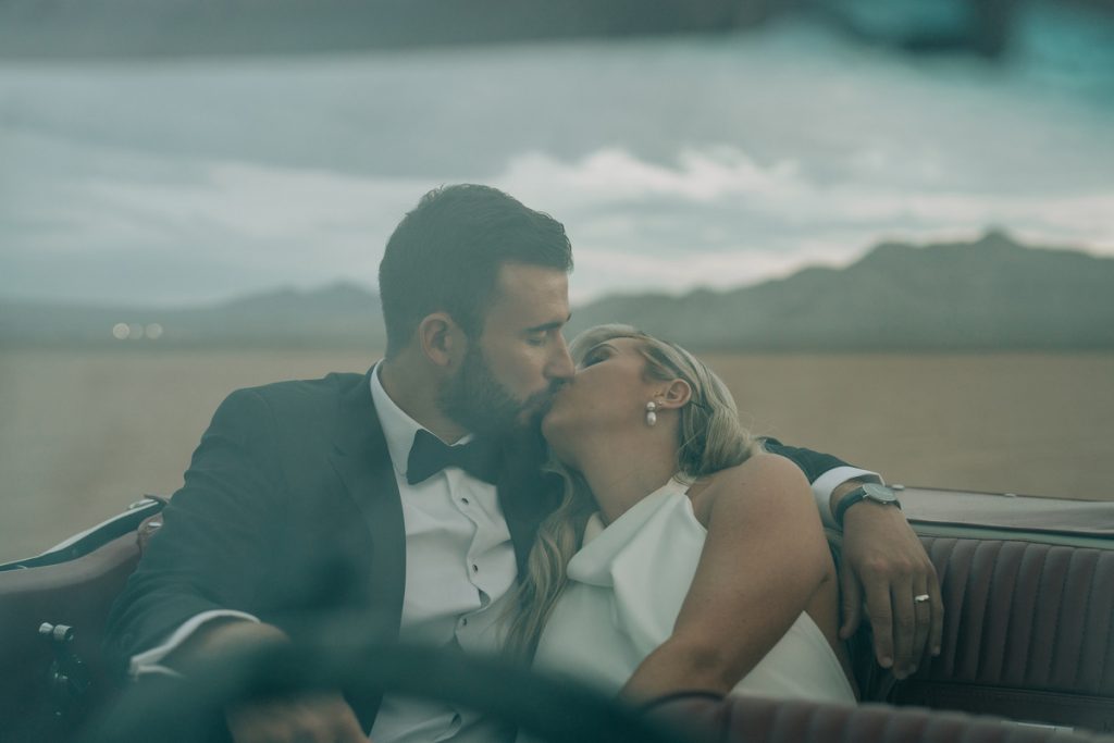 A bride and groom sit in the back seat of a classic convertible Mustang car and kiss. The Groom looks sharp in a suit with a bow tie, whilst the bride wears a minimalistic high neck white wedding dress.