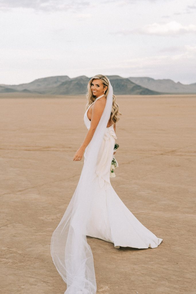 A bride stands in the middle of a desert during her Las Vegas wedding wearing a high neck minimal wedding dress with a thigh high split and large bow detailing on the back. She looks elegant and sophisticated with her blonde hair in Hollywood waves.