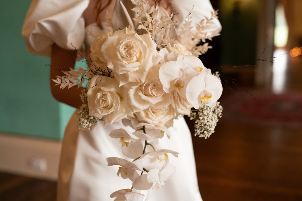 A large all white bouquet featuring roses, orchids and baby's breath being held by a bride wearing a white wedding dress with short puffy sleeves.
