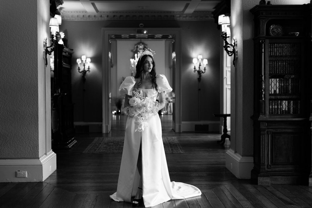 A regal looking bride stands within a corridor of a palatial manor, with wall lights illuminating her face. She wears a white wedding dress with a front split and the sleeves are short and puffy. She accessorises with a dramatic headpiece and an exquisite bouquet of delicate flowers.