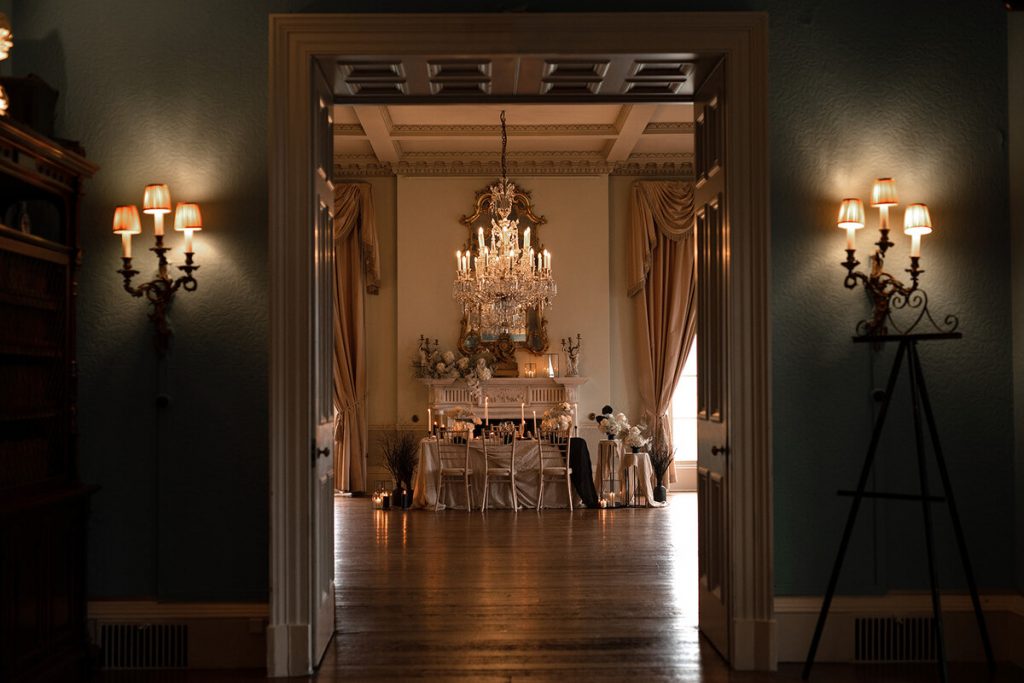 An intimate styled wedding tablescape is revealed set within a grand dining room of a stately home as the large doors are opened. The dinign table is styled with a colour palette of creams, stone tones and hints of black with a contemporary design led feel with lots of flickering lit candles decorating the table. The small tablescape features 6 cream wooden chairs and sits directly underneath a large dramatic chandelier.