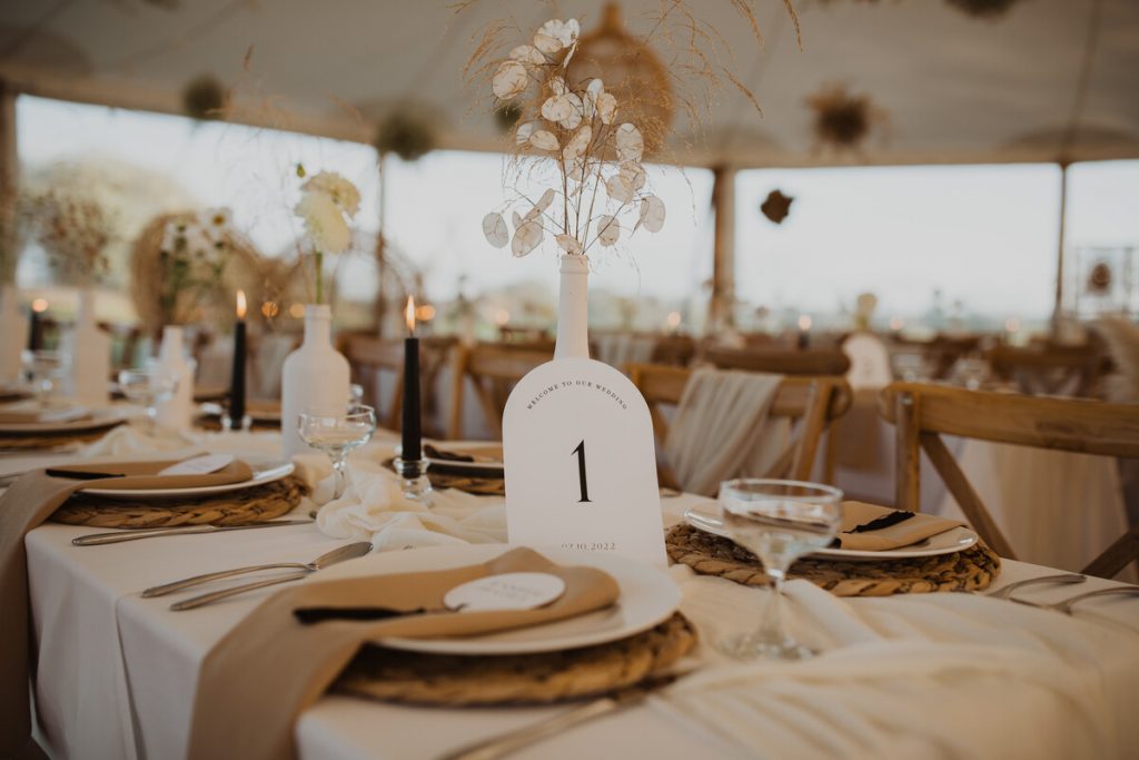 Long wedding tables clothed in beige fabric are dressed in a modern bohemian style with woven place mats and a palette of beige and creams mixed with hints of black. The tables are fully styled within a sailcloth tent.