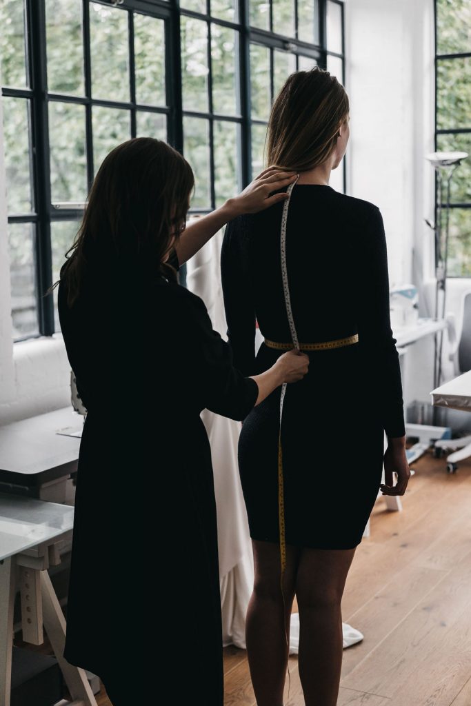 A bridal designer takes the measurements of her bride using a measuring tape with a view to making a bespoke bridal garment in her design studio