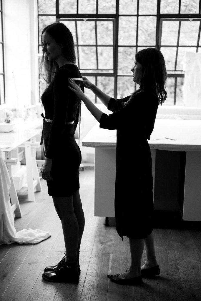 A bridal designer takes the measurements of her bride using a measuring tape with a view to making a bespoke bridal garment in her design studio