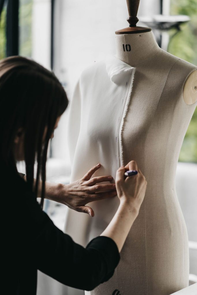 A bridal designer marks with pen measurements onto a cotton toile pinned to a mannequin whilst she makes a wedding dress.