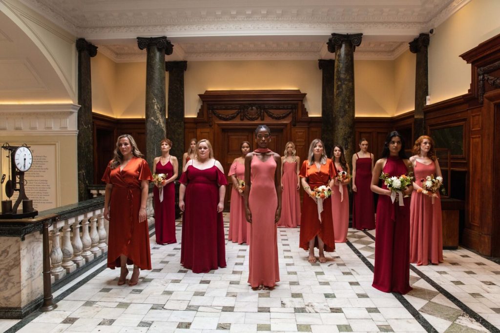 A large group of bridesmaids stand in a formation within a grand room with large pillars and high ceilings. The women wear mismatched bridesmaid dresses in various styles and differing shades of reds, pinks and burnt orange.