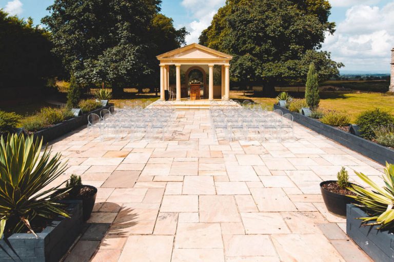 Clear ghost chairs are laid out neatly in rows with an empty aisle space facing a temple building ready for an outdoor wedding ceremony. Behind the temple are enormous green hedges and the skies above a wonderful shade of bright blue.