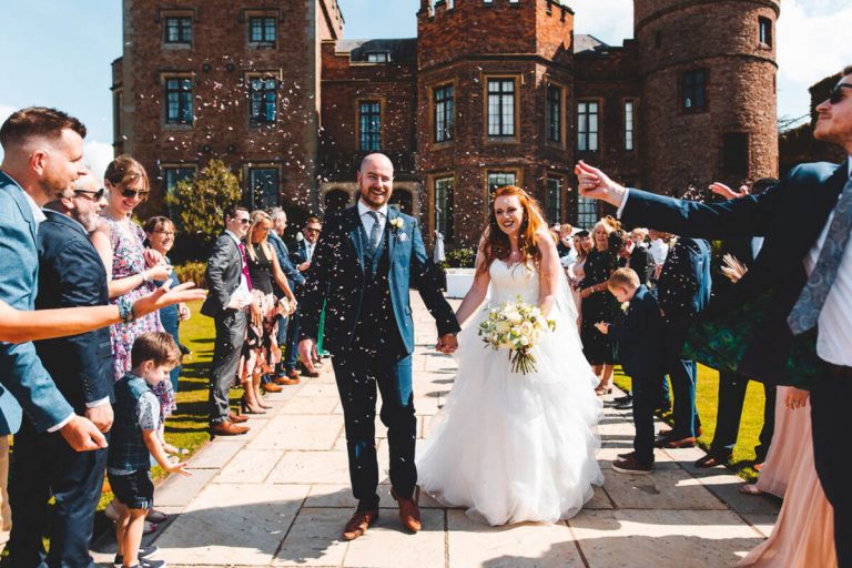 A bride and groom hold hands as they walk together between their group of guests who throw confetti over them. The newlyweds smile with joy. Behind them is a castle wedding venue.