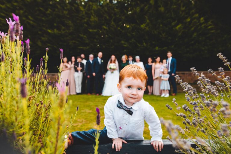 A gorgeous little brown haired toddler climbs up into the flower beds during group family photo. The family can be seen standing in the background.