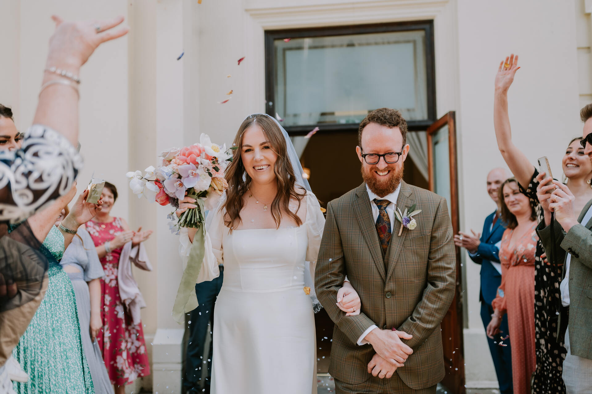 A newly married bride and groom walk arm in arm down the outside stairs of their ceremony space with huge beaming smiles of happiness as they are showered with colourful confetti from guests who stand either side of them on the steps.