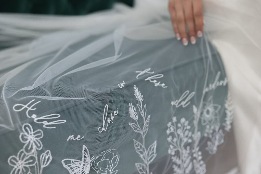 A hand embroidered veil lays on top of a green sofa with the words 'hold me close' sewn flawlessly into the sheer material accompanied by some floral detailing.