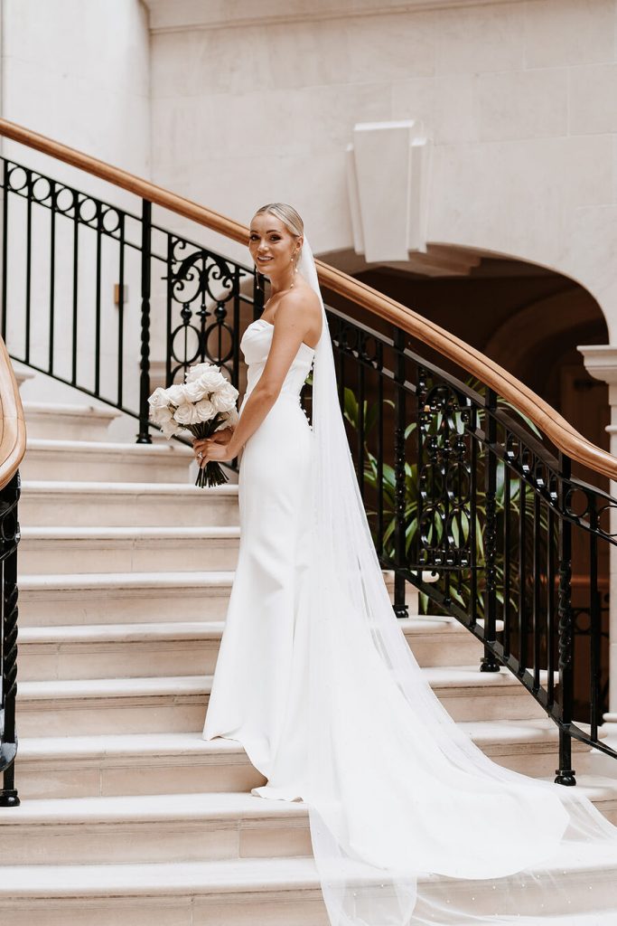 A bride wears a white strapless minimal wedding gown with a sweetheart neckline. Her blonde hair is tied in a low bun over which a long pearl veil is worn. She holds a large bouquet of fluffy white roses.