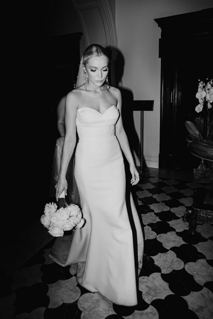 A bride wears a white strapless minimal wedding gown with a sweetheart neckline. Her blonde hair is tied in a low bun over which a long pearl veil is worn. She holds a large bouquet of fluffy white roses.