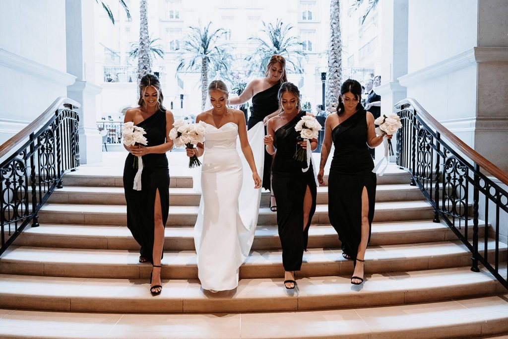 A bride wearing a white strapless wedding dress with a sweetheart neckline stands between her 4 bridesmaids who wear a one sleeved matching balck wedding dress. All the ladies hold a large bouquet of fluffy white roses.
