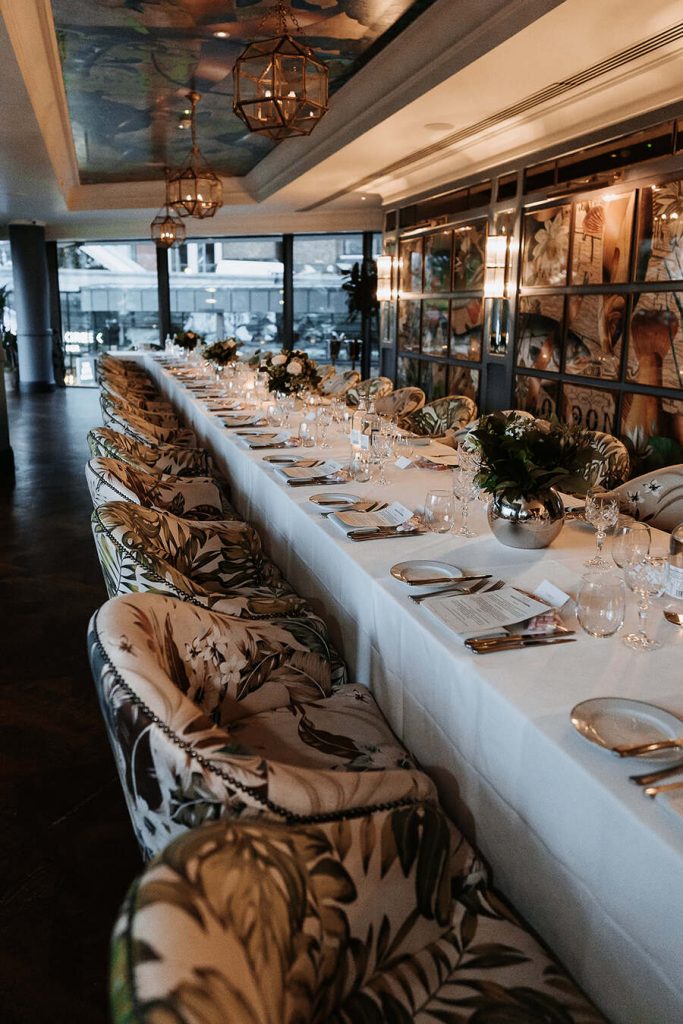 A private dining room is set with a long white clothed table around which comfortable tub chairs with a jungle theme print are set. The table is styled with minimal decor giving a sophisticated, elegant feel.