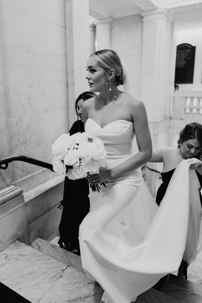 A bride wears a white strapless minimal wedding gown with a sweetheart neckline. Her blonde hair is tied in a low bun over which a long pearl veil is worn. She holds a large bouquet of fluffy white roses.