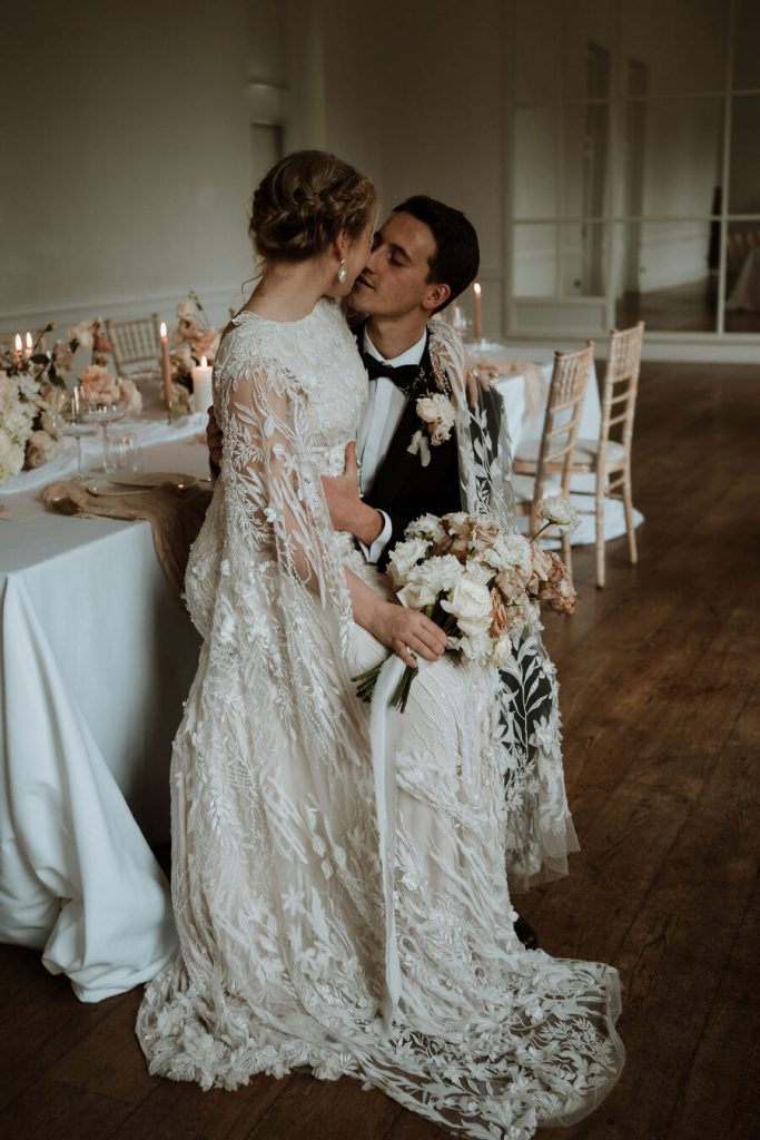 An elegant wedding dress features delicate, intricate hand beaded lace and a dramatic detailed cape.