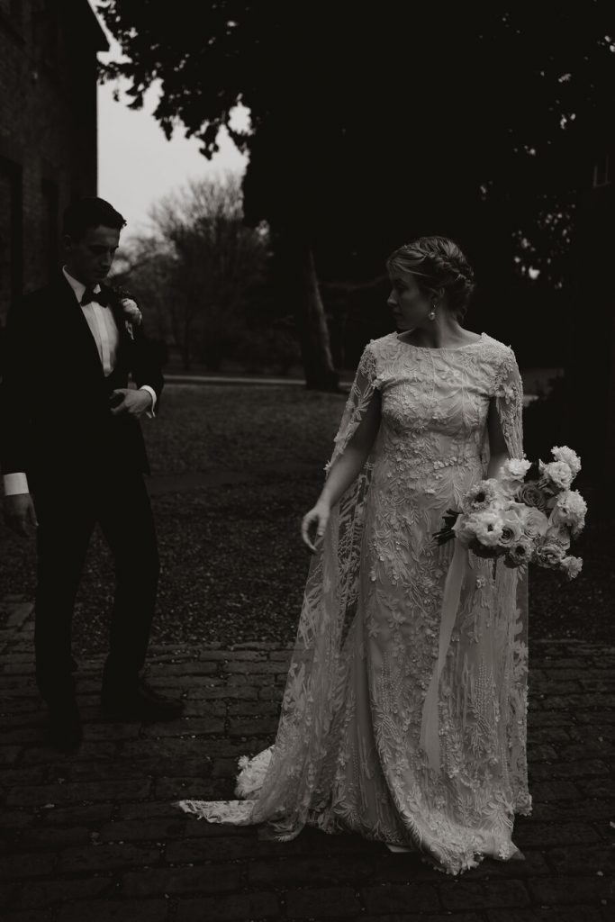 An elegant wedding dress features delicate, intricate hand beaded lace and a dramatic detailed cape.