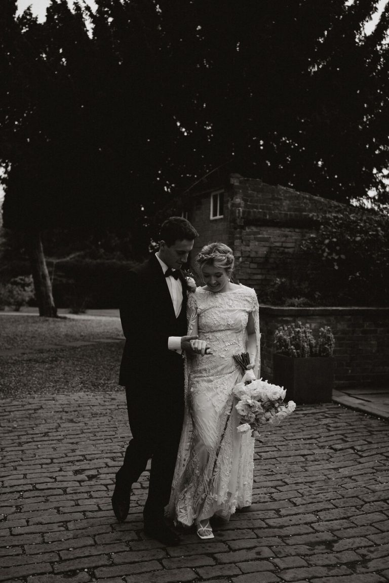 A married couple stroll through the grand grounds of their wedding venue, alone in their own newly married bubble. The groom looks smart wearing a tuxedo suit and bow tie whilst the bride wears a delicate lace beaded wedding dress with cape detailing and her hair in a braided halo up do.