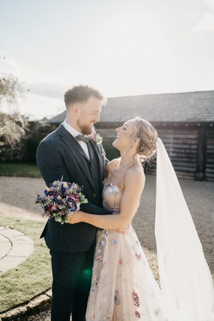 A bride a groom wander their wedding venue's grounds for couple portraits. The groom wears a smart blue woollen suit with a bow tie whilst his bride wears a sweetheart neckline colourful hand painted wedding dress.