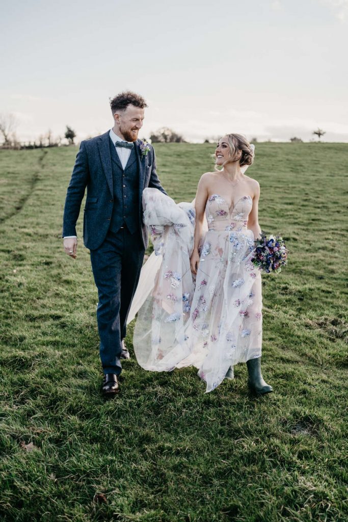 A bride a groom wander their wedding venue's grounds for couple portraits. The groom wears a smart blue woollen suit with a bow tie whilst his bride wears a sweetheart neckline colourful hand painted wedding dress.