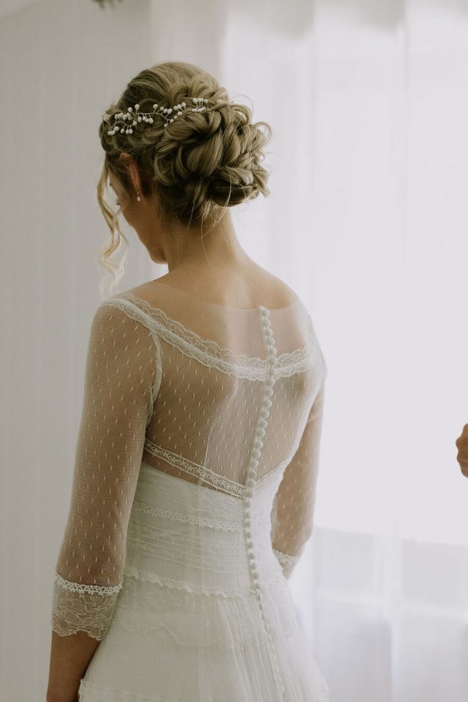 An untraditional bride wears a vintage style wedding dress with a sheer overlay detailed top and her hair worn up with a freshwater pearl hair accessory and freshwater pearl drop earrings.