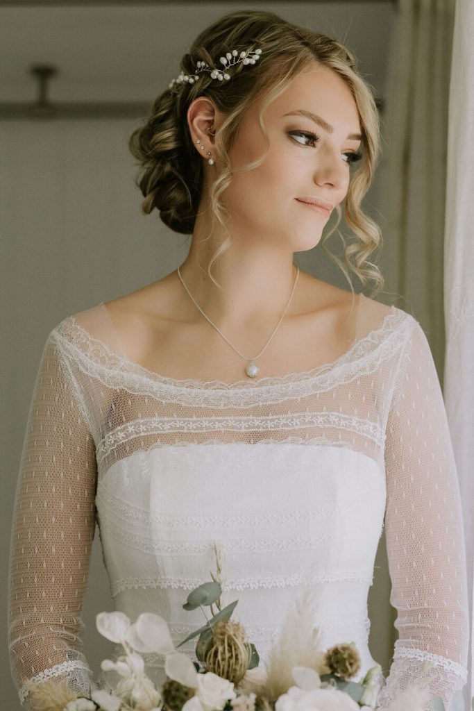 An untraditional bride wears a vintage style wedding dress with a sheer overlay detailed top and her hair worn up with a freshwater pearl hair accessory and freshwater pearl drop earrings.