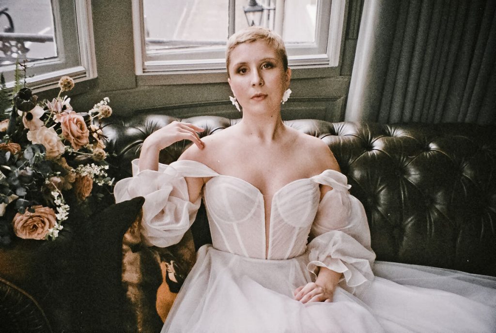 Close up editorial bridal frame of a bride with super short bleached hair, wearing a sweetheart neckline floaty blush coloured wedding dress, sitting on a large leather Chesterfield sofa looking straight down the camera. The image is shot on film, giving a grainy, textured aesthetic to the image.