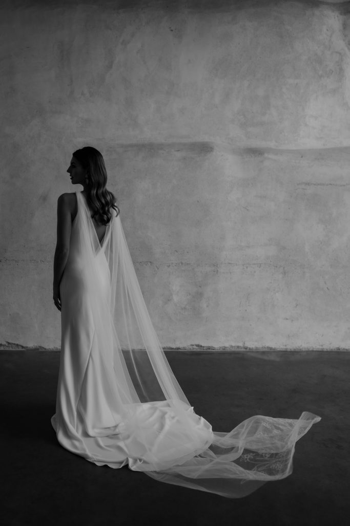 An alternative to a traditional wedding veil; an embroidered wedding cape is attached to the shoulders of a wedding dress, draping gently down to the floor where stitched florals and writing are embellished.