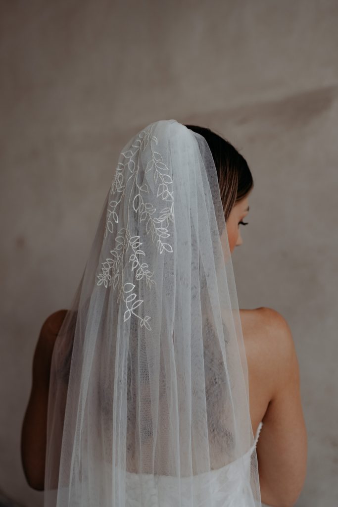 A beautiful bride with long brunette hair wears a long sheer wedding veil with ruscus flowers embroidered along the entire length of the hem.