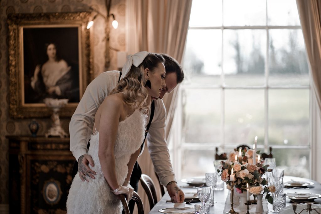 A bride and groom look lovingly at their wedding table fully set up prior to guests arriving. The table is laid with footed bowls of flowers interspersed with lit candles and has a 1930s vintage style. The groom stands behind his wife, holding her gently around the waist. The wife has a 1930s vintage look with sheer pearl studded gloves, a large white hair bow and tight Hollywood curls.