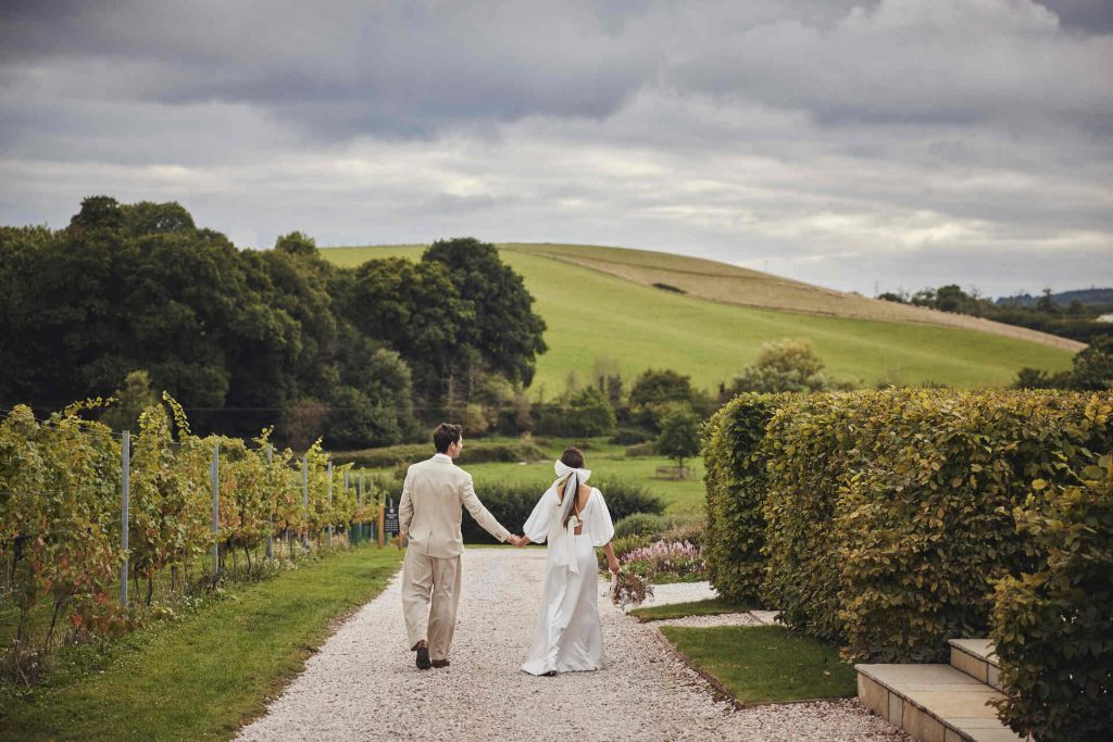 A quiet stroll around the estate of Devonshire's Brickhouse Vineyard for a newlywed couple. The rolling green countryside hills and woodlands can be seen in the distance with beautiful hedgerows and small trees lining the path where the couple walk hand in hand.