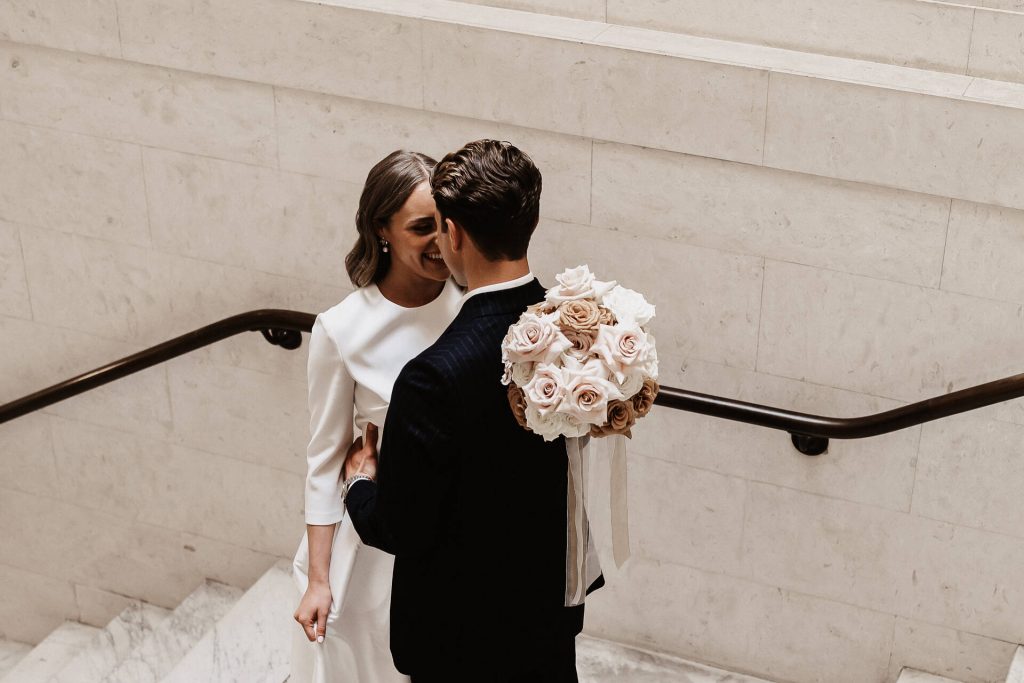A bride and groom share a quiet moment together on the steps of their wedding venue. They gently embrace, the bride putting one hand over her husband's shoulder whilst she holds a beautiful bouquet of roses.