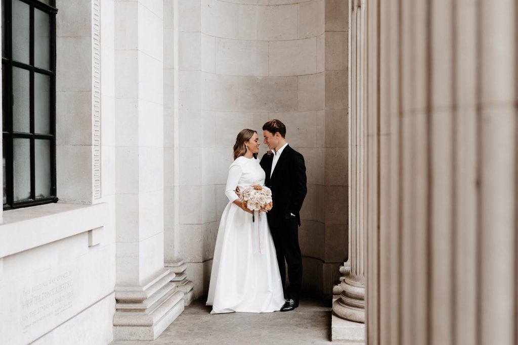 A newly wed couple stand in the iconic stone walled alcove beyond the Corinthian columns at London's iconic Old Marylebone Town Hall