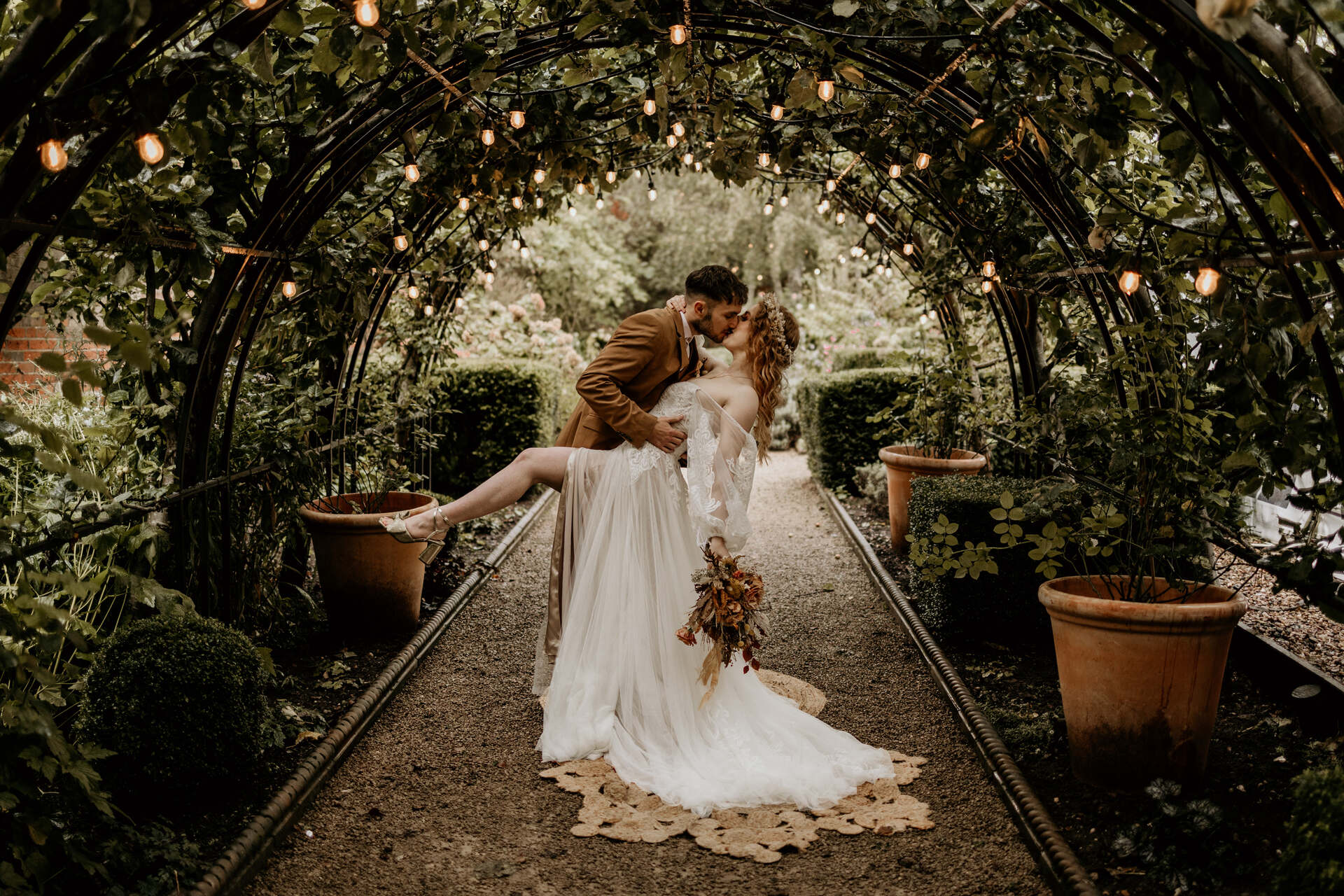 A bride and groom dip for a kiss underneath a tunnel of greenery and lights. The bride, who wears a flowing boho wedding dress leans far back and holds one bare leg high up in the air. The scene is beautifully romantic.