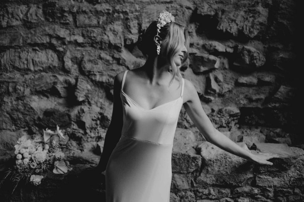 A blonde bride wears a minimalist white slip dress with her hair worn up and loose waves cascading down the front of her face. She wears a dramatically embellished 3d floral headband. An elegant, soft look against the exposed distressed wall of a historic building behind her.