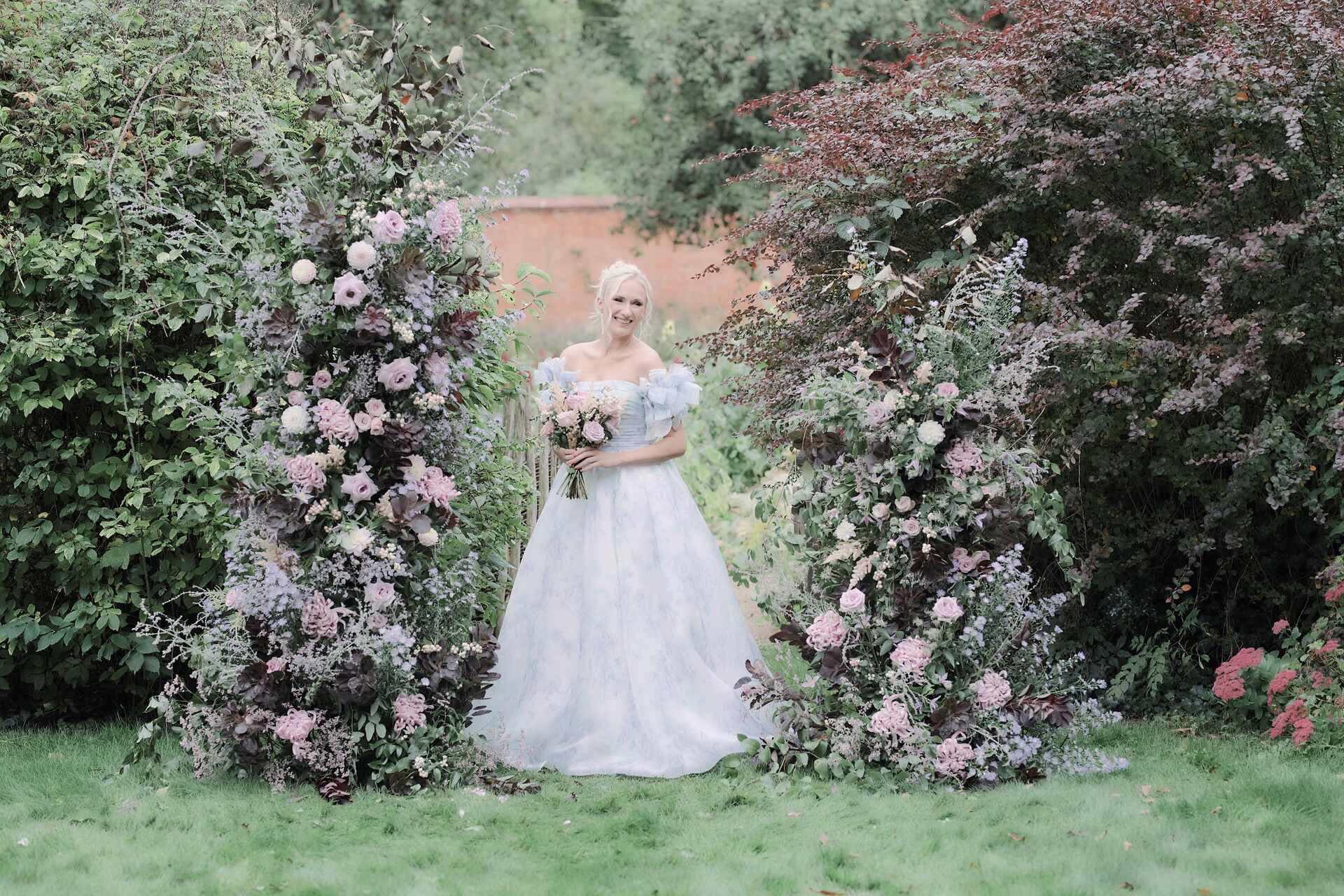 A bride wearing an off the shoulder floaty wedding dress with hints of blue is framed by an asymmetric broken floral arch with a Victorian walled garden as a romantic, English countryside wedding backdrop.
