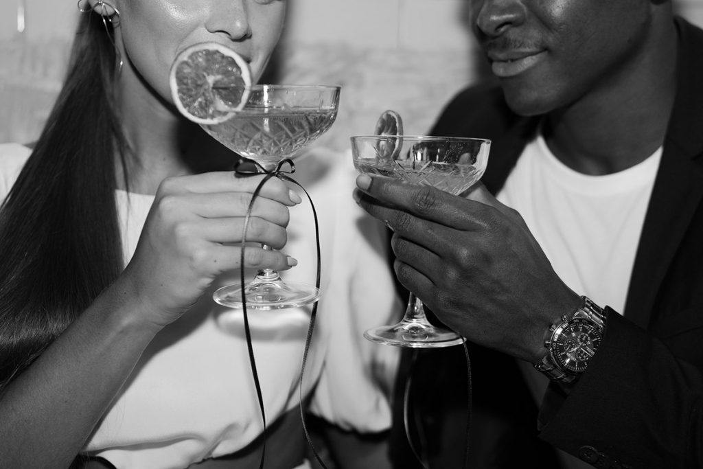 A couple cheers one another with cut crystal Champagne coupe glasses. The glasses have a slice of dried fruit decorating the rim with a delicate thin bow ribbon embellishing the stem of the glass for a contemporary, design-led wedding aesthetic
