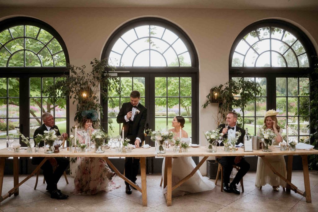 A groom stands holding a microphone mid speech at his wedding. His wife sits next to him at the wooden top table, gazing in adoration at her husband. Other close family members sit wiping tears of joy and emotion from their faces as the groom speaks..
